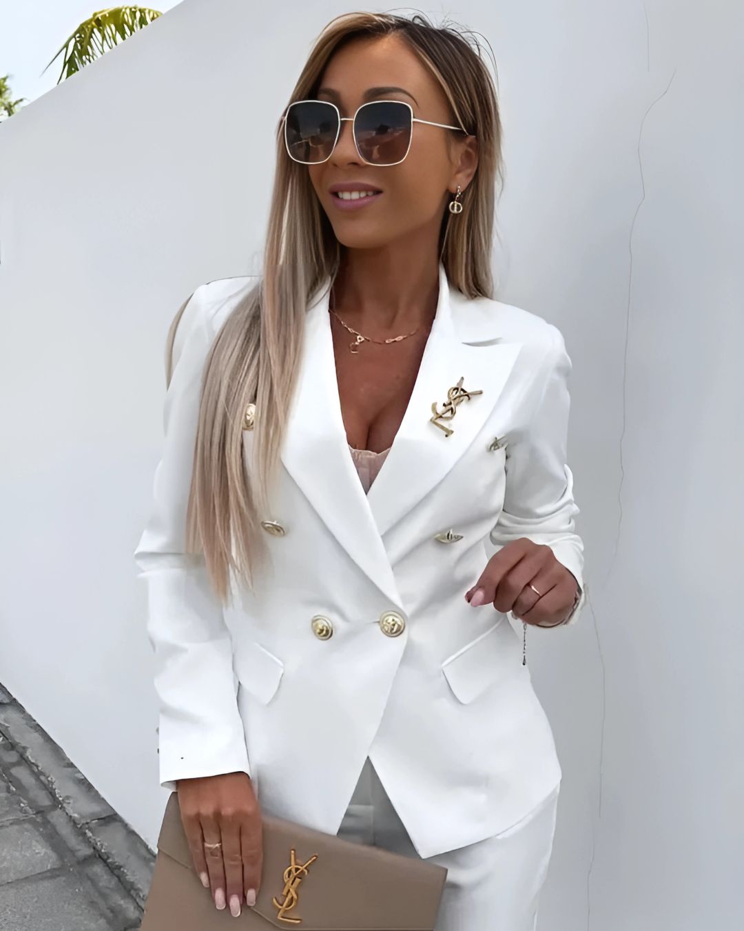 Frau in weißem Blazer mit goldenen Knöpfen und YSL-Logo, trägt Sonnenbrille und hält eine beige YSL-Clutch. Elegante Mode, Luxus-Accessoires.