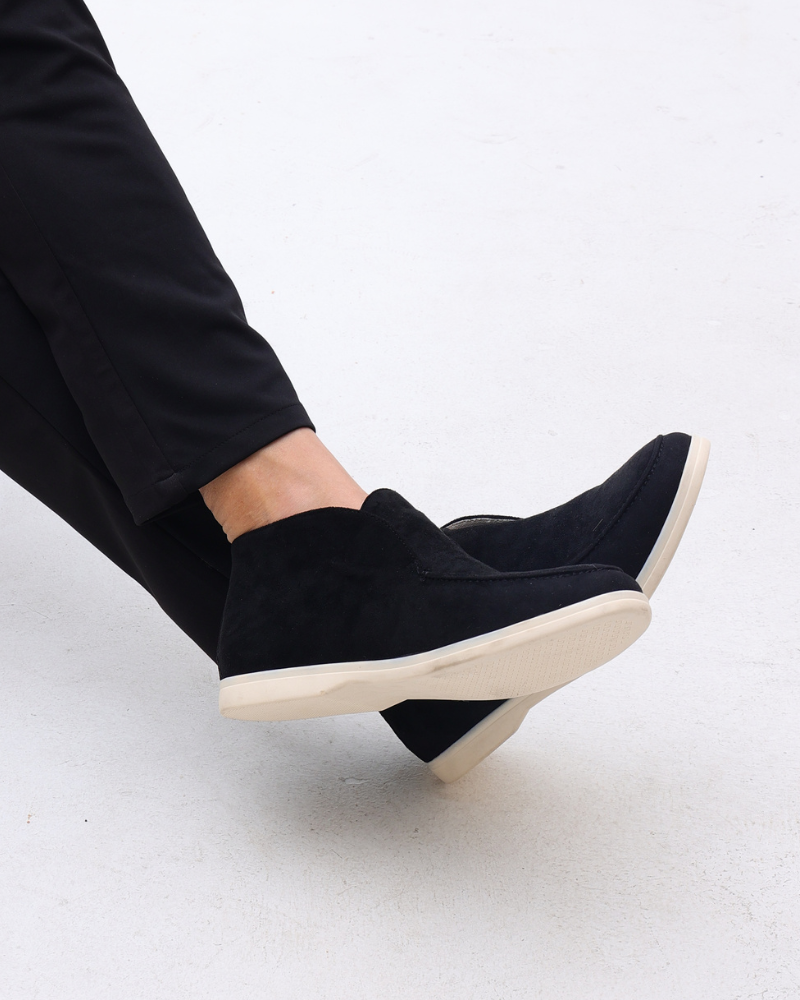 Schwarze Wildleder-Sneaker mit weißer Sohle, getragen von einer Person in schwarzer Hose, auf hellem Hintergrund. Elegante, moderne Freizeitschuhe.