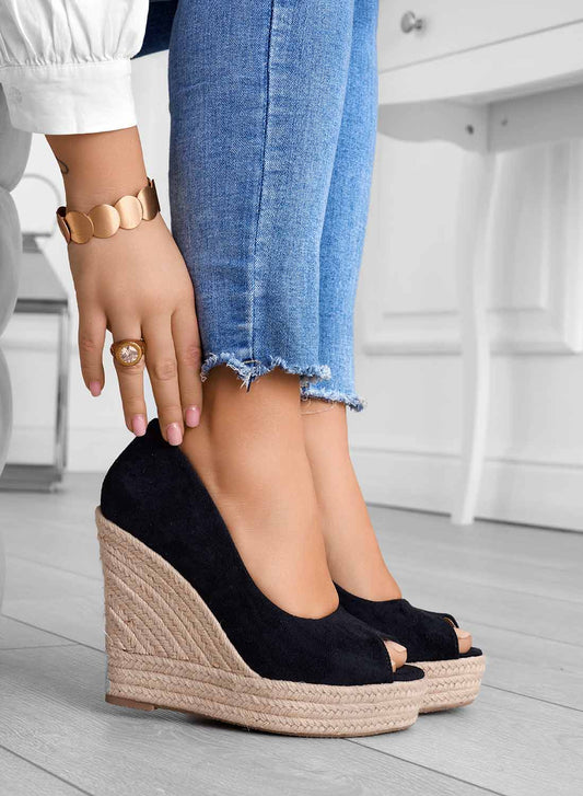 Damenfüße in blauen Jeans und schwarzen Keilabsatz-Sandalen aus Wildleder auf Holzboden. Modischer Schmuck am Handgelenk.