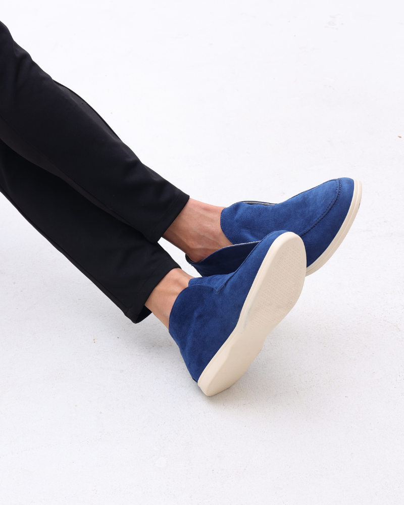 Blaue Wildleder-Slipper an den Füßen einer Person in schwarzen Hosen, auf hellem Hintergrund. Bequeme, stilvolle Freizeitschuhe für Herren.