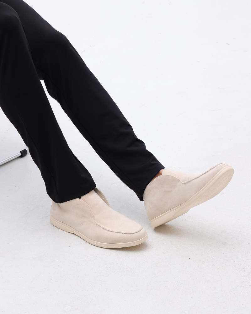 Beige Wildleder-Slipper an Füßen einer sitzenden Person in schwarzer Hose auf weißem Hintergrund. Elegante, bequeme Herrenschuhe.