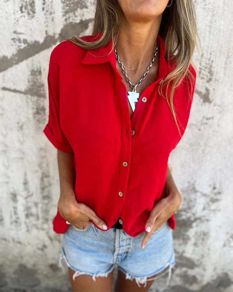 Frau in roter Bluse und Jeansshorts vor Betonwand, modischer Sommerlook, lässiger Stil, silberne Halskette, trendige Damenmode.