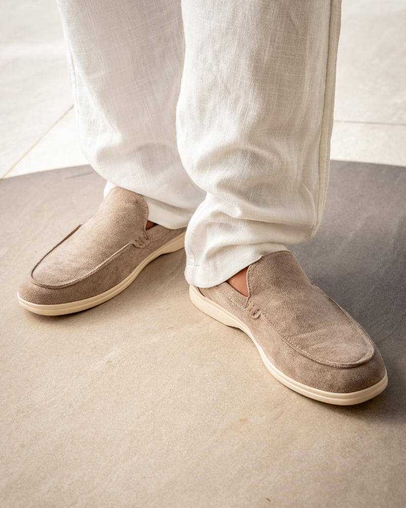 Beige Wildleder-Slipper auf grauem Boden, getragen mit weißen Leinenhosen. Elegante Herrenschuhe, ideal für Sommermode und Freizeitkleidung.