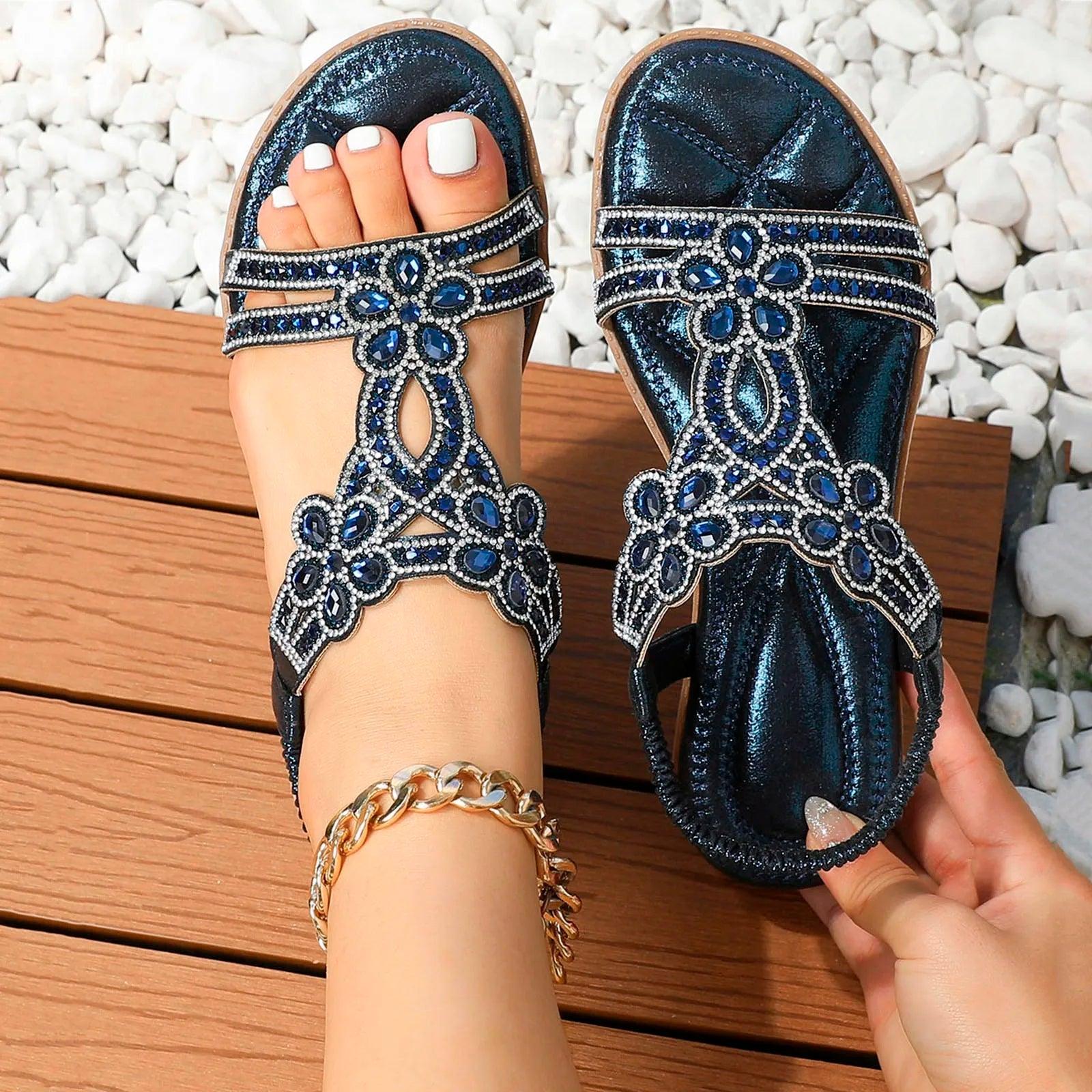 Blaue Sandalen mit Strasssteinen auf Holzterrasse, weiße Pediküre, goldene Fußkette, modische Sommerschuhe, elegantes Design.
