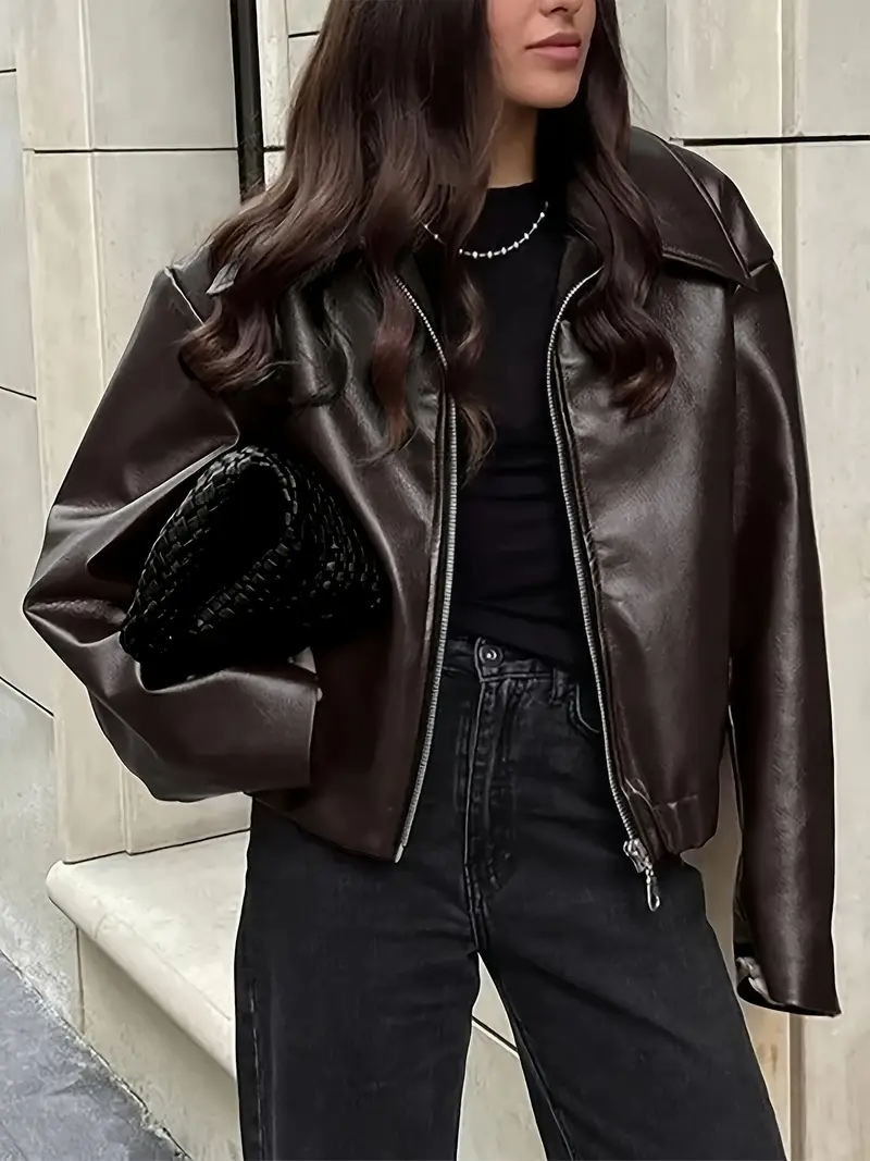 Frau in brauner Lederjacke, schwarzem Top und Jeans, hält schwarze geflochtene Tasche. Modetrend Herbst, Streetstyle, elegante Damenmode.