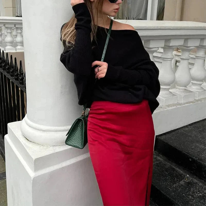 Frau in schwarzem Pullover und rotem Rock lehnt an weißer Balustrade, trägt grüne Handtasche. Mode, Streetstyle, elegantes Outfit.