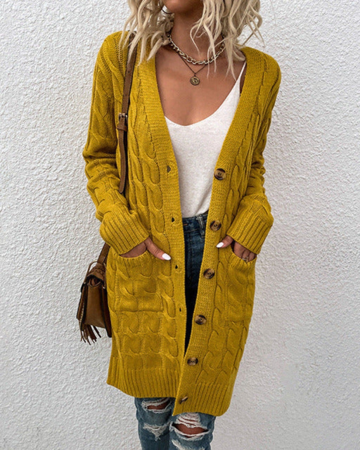 Frau in gelbem Strickcardigan mit Zopfmuster, weißem Top und zerrissenen Jeans vor weißer Wand. Modetrend, Herbstmode, lässiger Stil.