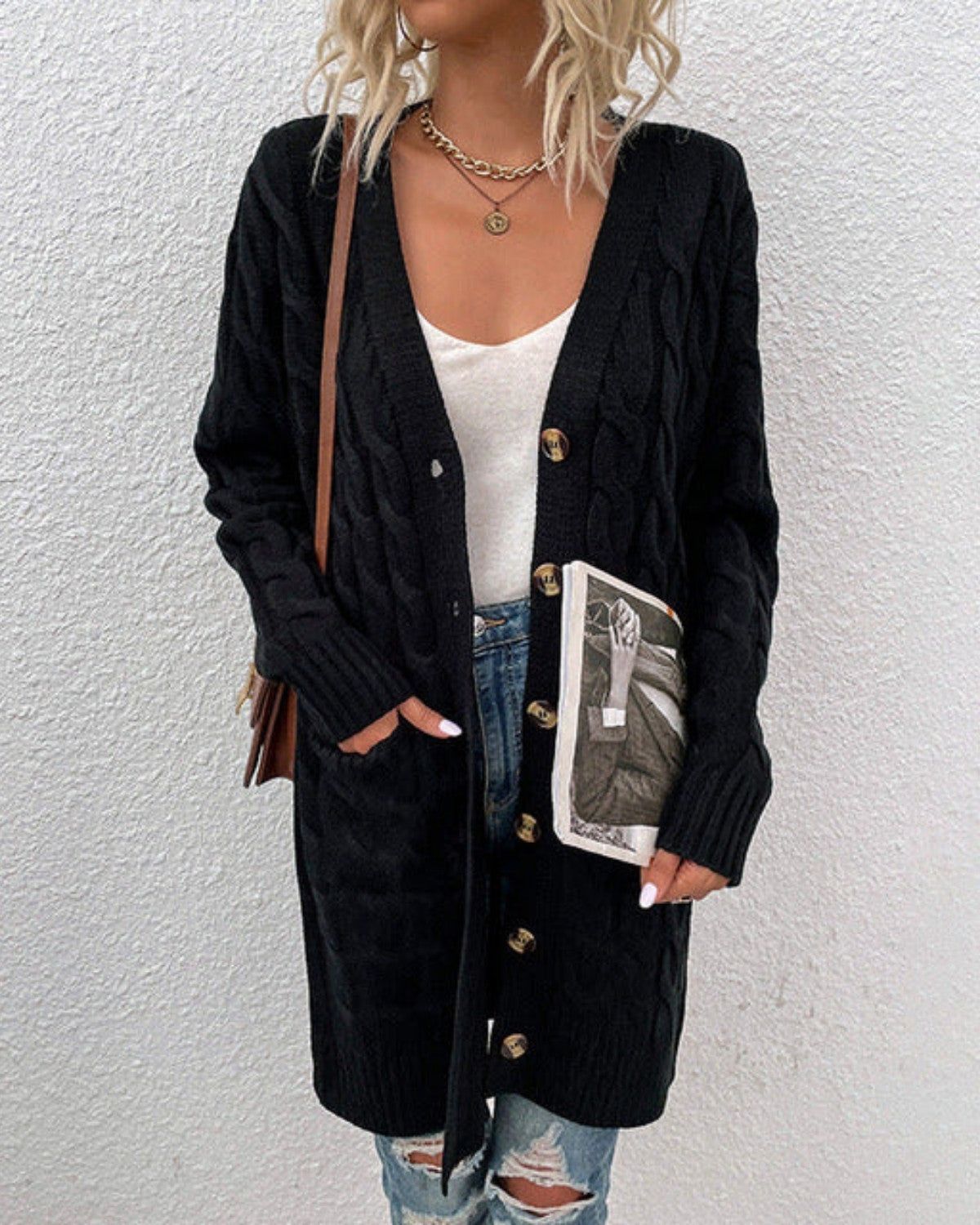 Frau in schwarzer Strickjacke mit Zopfmuster, weißem Top und zerrissenen Jeans, hält ein Buch. Modetrend, lässiger Herbstlook, Damenmode.