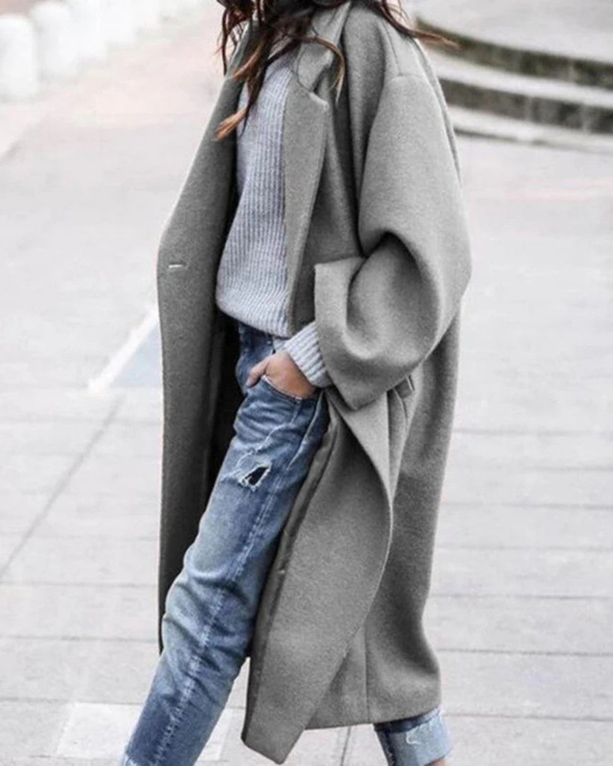 Frau in grauem Oversize-Mantel, grauem Pullover und zerrissenen Jeans spaziert draußen. Modetrend, Herbstmode, lässiger Stil.