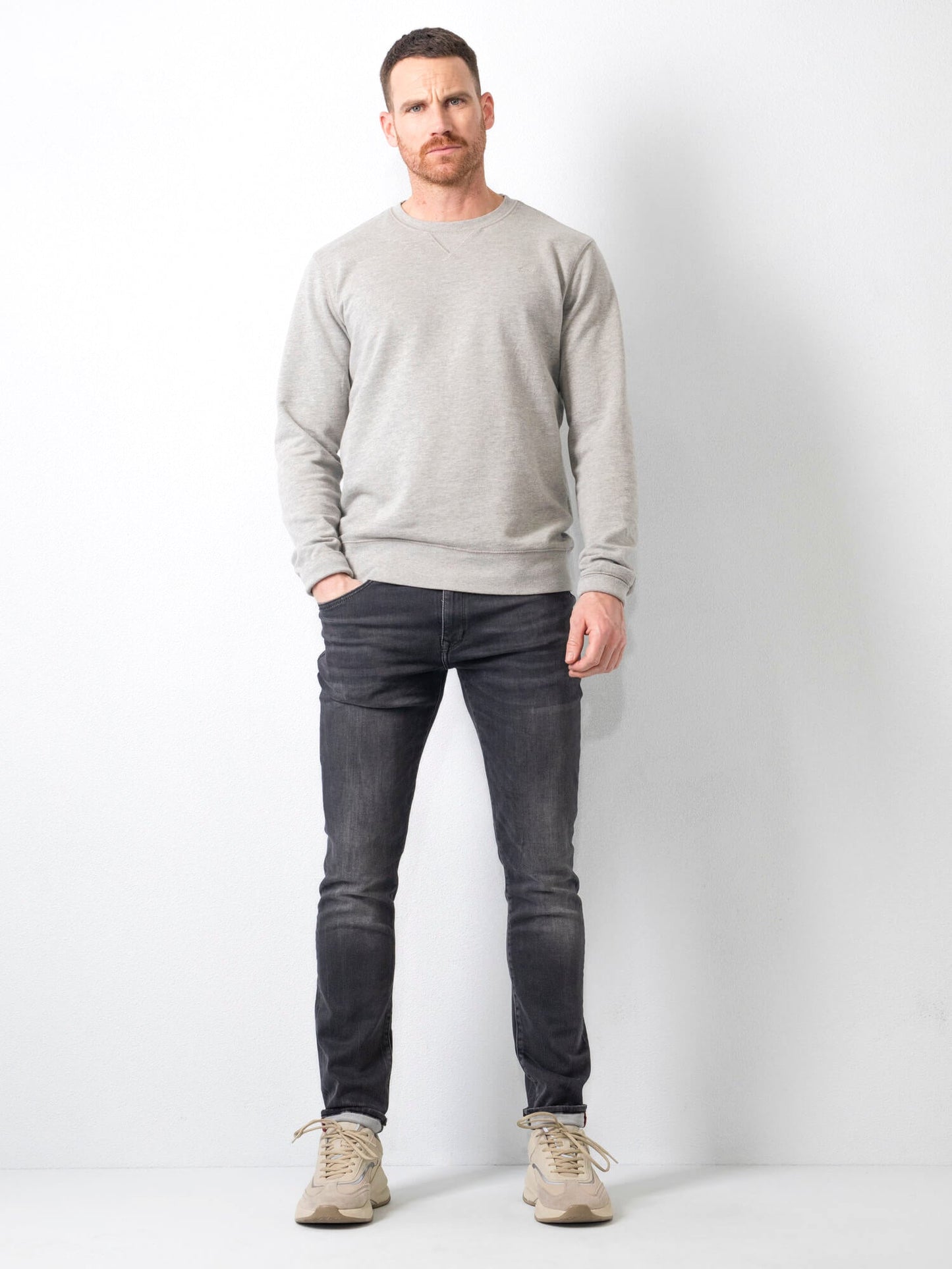Mann in grauem Pullover und dunklen Jeans vor weißem Hintergrund, lässiger Look, Mode für Herren, Freizeitkleidung, moderne Herrenmode.