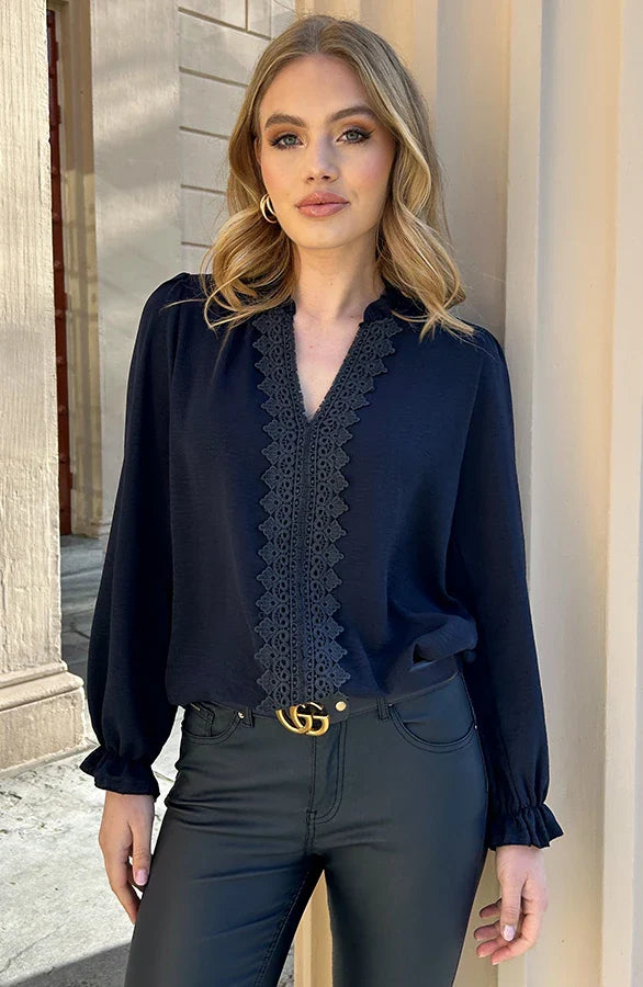 Frau in eleganter schwarzer Bluse mit Spitzenbesatz und Lederhose, steht vor einem Gebäude. Mode, Damenbekleidung, stilvoller Look.