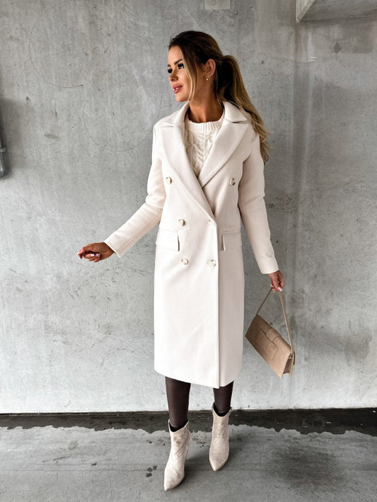 Frau in elegantem, weißem Wollmantel, kombiniert mit beiger Handtasche und Stiefeletten, vor grauer Betonwand. Wintermode, stilvoll, modern.