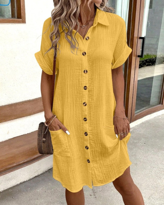 Frau in gelbem Leinenkleid mit Knopfleiste, kurzen Ärmeln und Taschen, steht vor einer Bank. Sommermode, lässiger Stil, Damenbekleidung.