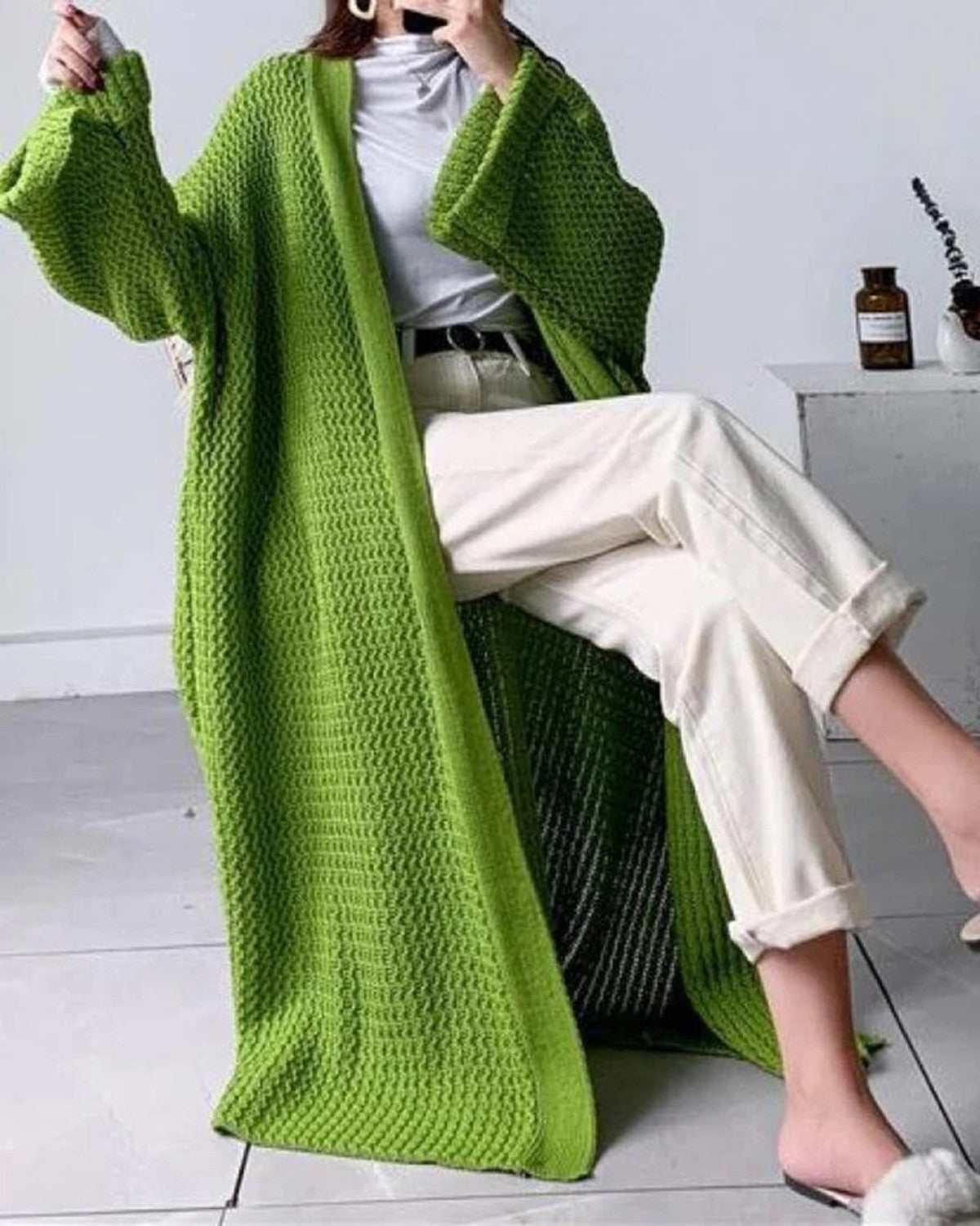 Frau in grünem Strickmantel, weißem Rollkragenpullover und beigen Hosen sitzt lässig. Modetrend, Strickmode, Herbstoutfit.