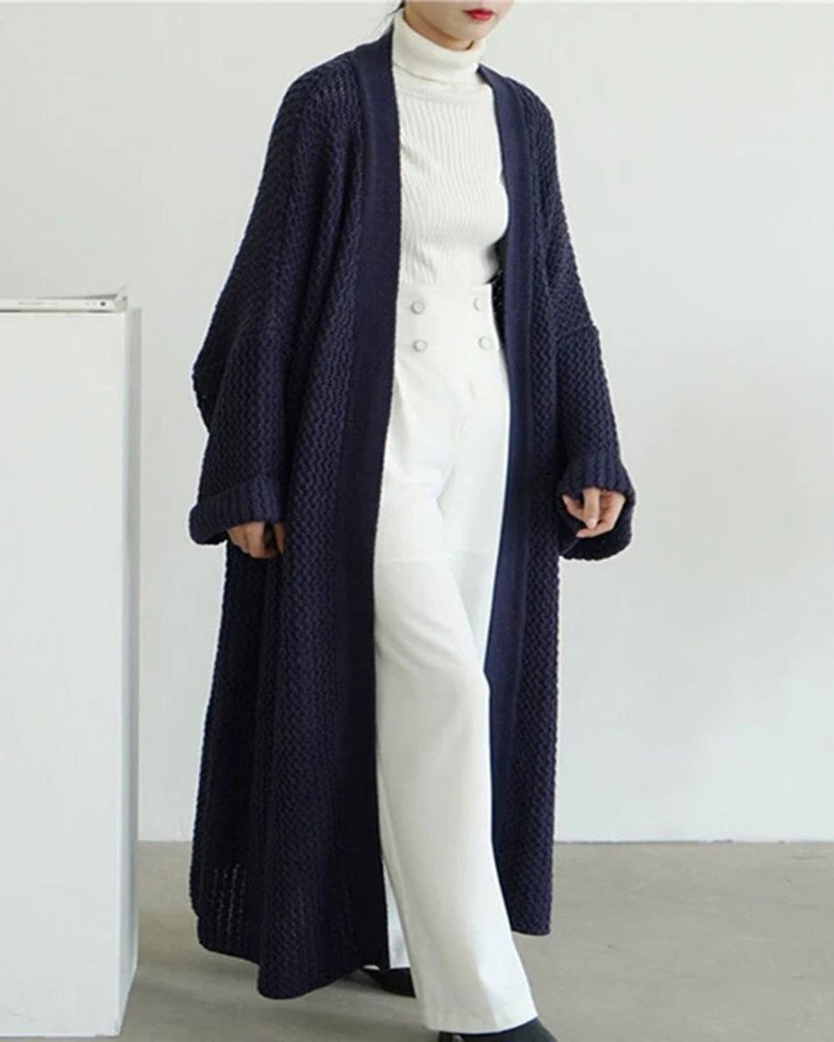 Dame in weißem Rollkragenpullover und Hose, trägt langen, dunkelblauen Strick-Cardigan. Modetrend, eleganter Herbst-Look, minimalistisch.