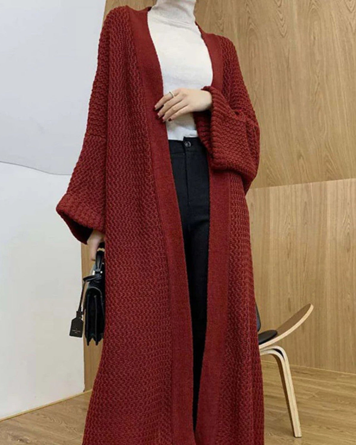 Frau in langem, rotem Strick-Cardigan, weißem Rollkragenpullover und schwarzer Hose vor Holzwand. Modetrend, Herbstmode, Damenbekleidung.