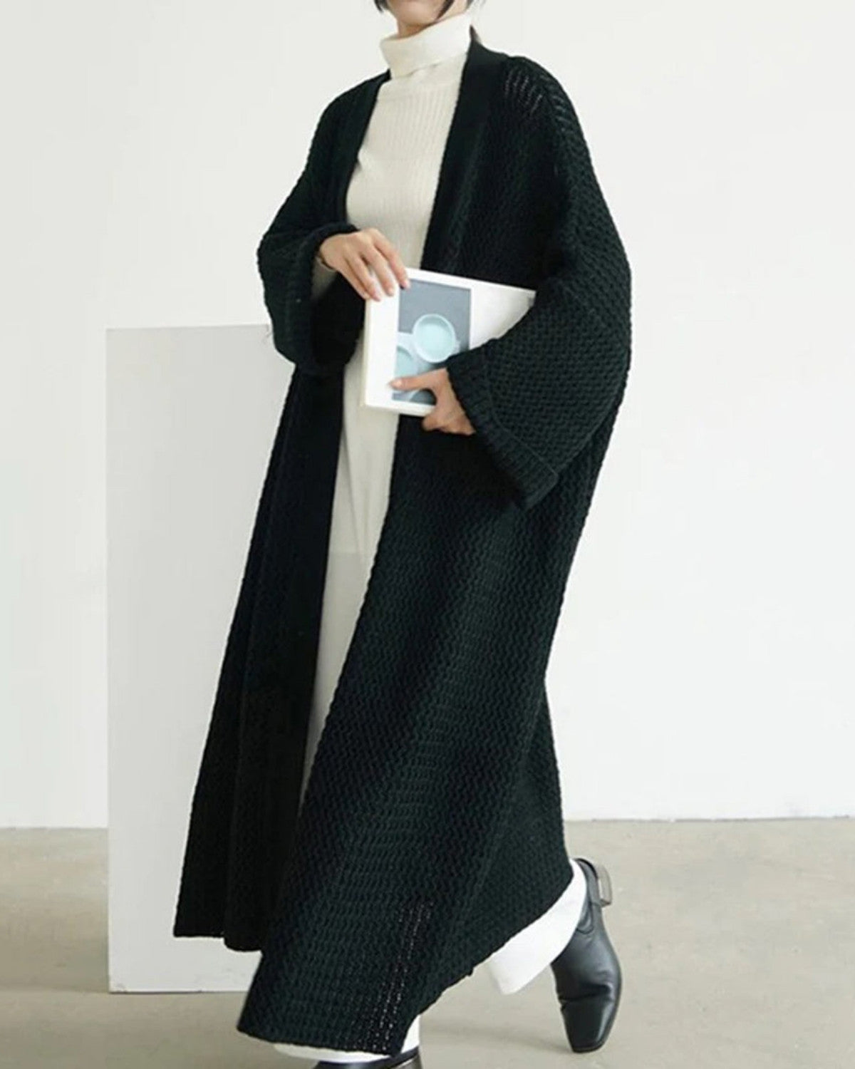 Frau in langem, schwarzem Strickmantel und weißem Rollkragenkleid hält ein Buch. Modetrend, Herbstmode, eleganter Look, minimalistischer Stil.