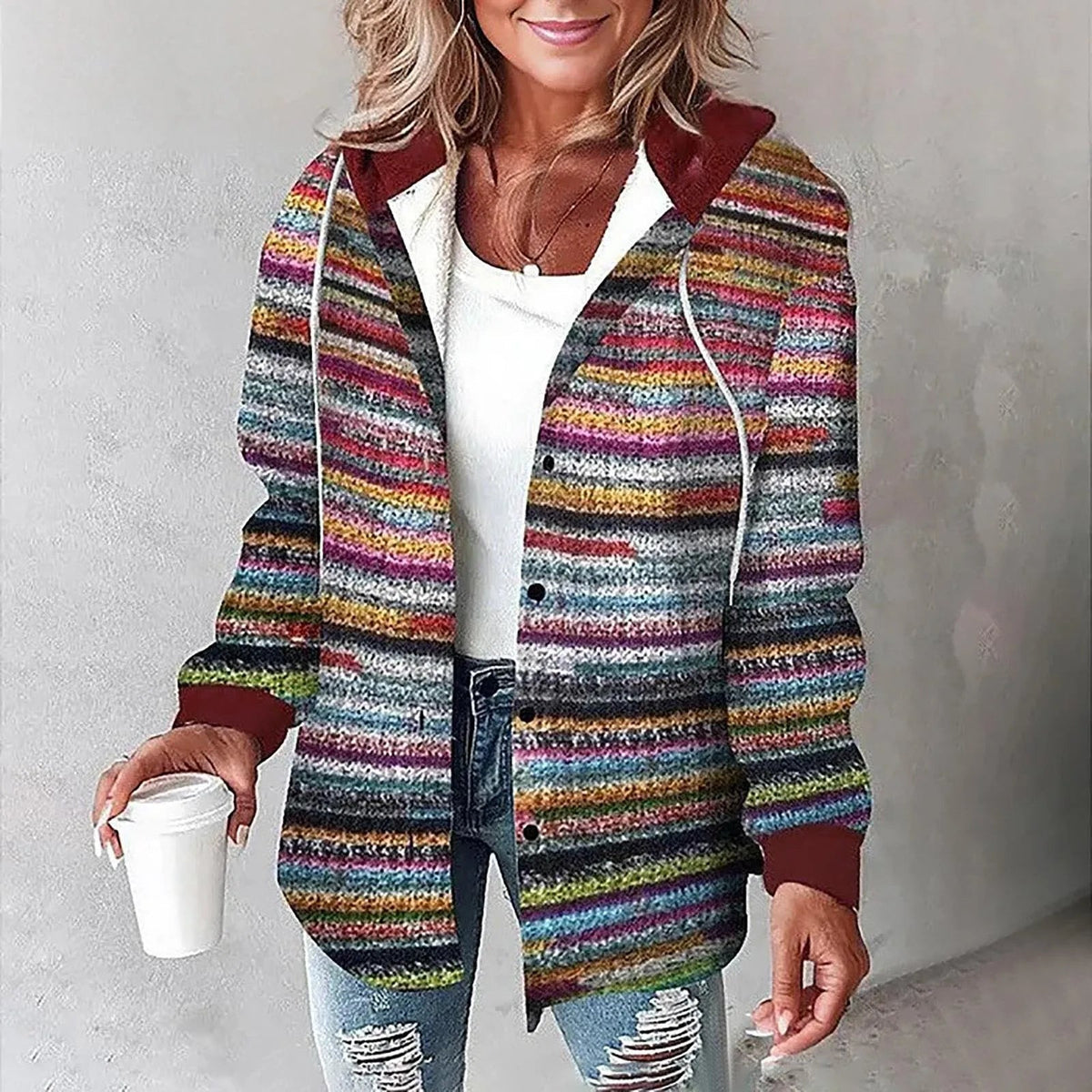 Frau in bunter, gestreifter Strickjacke mit Kapuze, hält weißen Kaffeebecher, trägt zerrissene Jeans. Modetrend, Herbstmode, lässiger Stil.