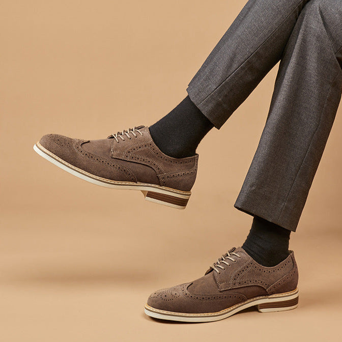 Braune Herren-Brogues aus Wildleder, kombiniert mit grauer Anzughose und schwarzen Socken, auf beigem Hintergrund. Elegante Herrenschuhe.