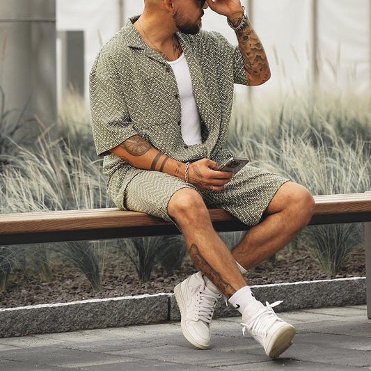 Mann in gemustertem Sommeranzug sitzt auf Bank, hält Smartphone. Trendige Herrenmode, lässiger Stil, urbane Umgebung.