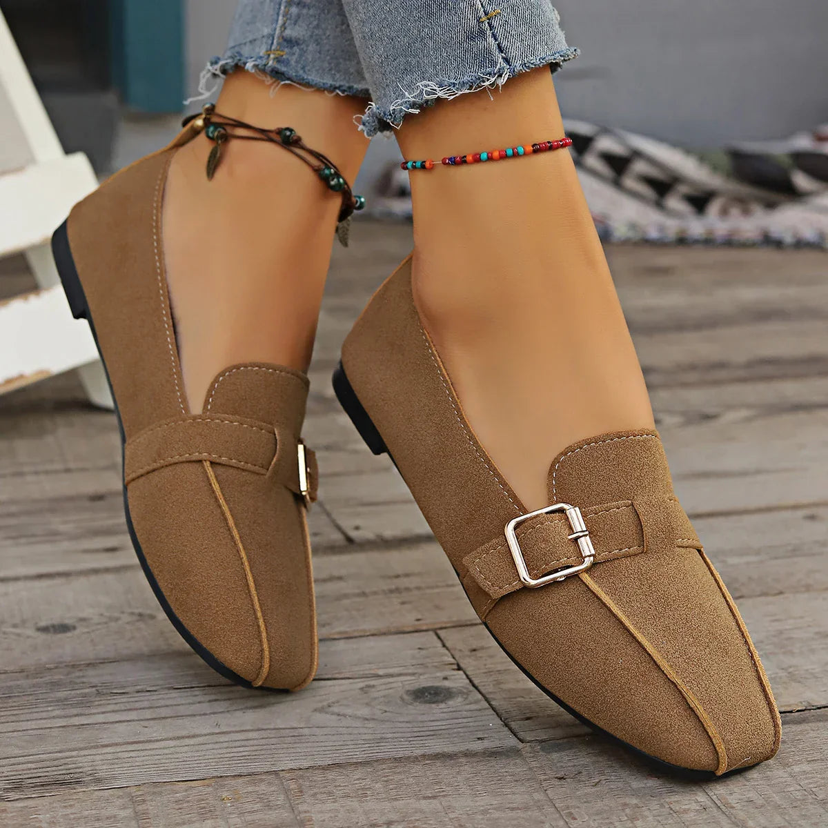 Braune Damen-Loafer mit Schnalle, getragen mit Jeans und bunten Fußkettchen, auf Holzboden. Modische, bequeme Schuhe für den Alltag.