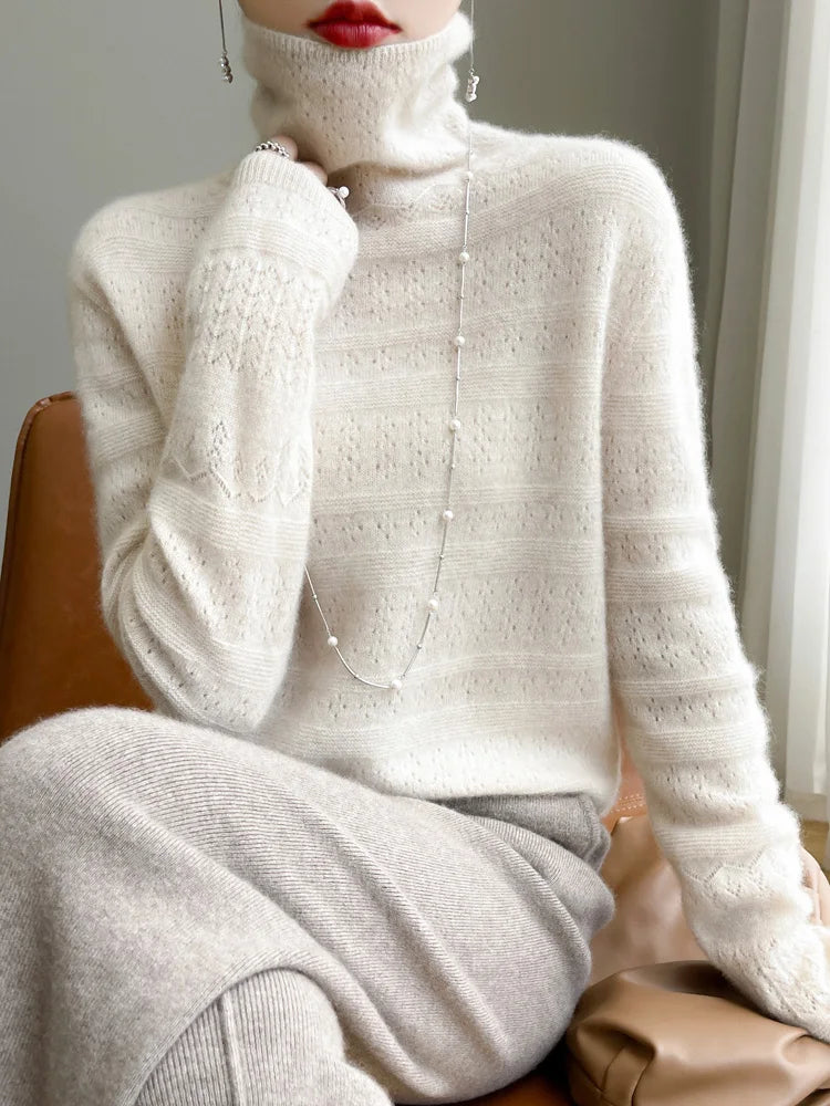 Frau in cremefarbenem Strickpullover mit Rollkragen, eleganter Halskette und rotem Lippenstift, sitzt auf braunem Stuhl. Modetrend Herbst/Winter.