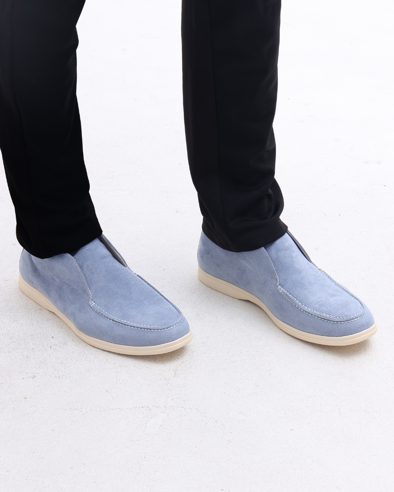 Herrenbeine in schwarzen Hosen mit hellblauen Wildleder-Slippern auf weißem Hintergrund. Elegante, bequeme Herrenschuhe, modisch, stilvoll.