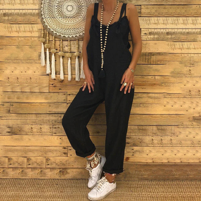 Frau in schwarzem Jumpsuit vor Holzwand, trägt weiße Sneakers und lange Perlenkette. Boho-Dekoration im Hintergrund. Modetrend, lässiger Stil.
