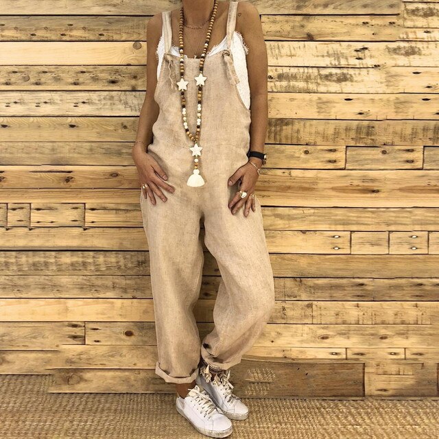 Frau in beiger Leinen-Latzhose vor Holzwand, trägt lange Halskette und weiße Sneakers. Modetrend, lässiger Stil, nachhaltige Kleidung.