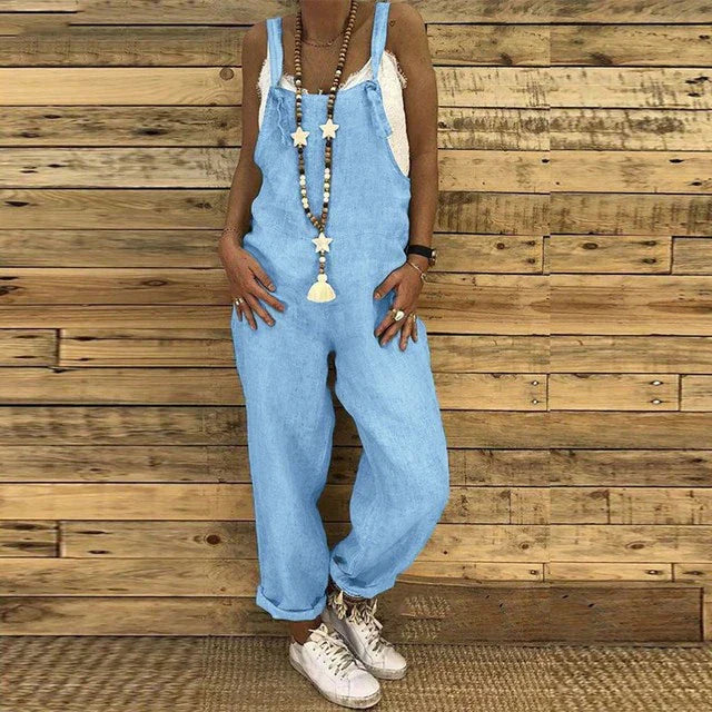 Frau in blauer Latzhose vor Holzwand, trägt weiße Sneakers und lange Halskette. Modetrend, lässiger Stil, Damenmode, Streetwear.
