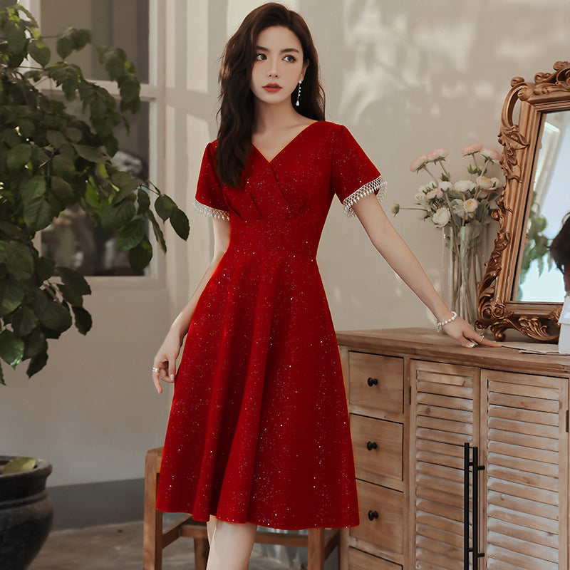 Frau in elegantem roten Kleid mit Glitzer, steht neben einer Kommode und einem Spiegel, umgeben von Pflanzen und Blumen, stilvolle Abendmode.