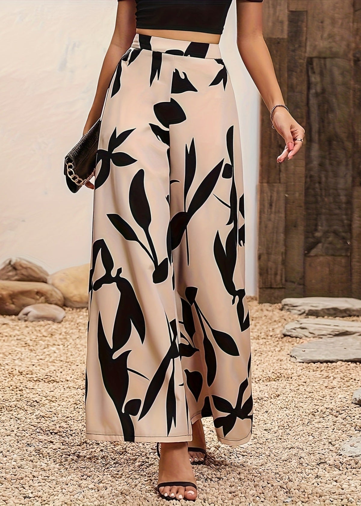 Frau in eleganter, hoch taillierter, schwarz-weiß gemusterter Palazzohose, kombiniert mit schwarzem Oberteil und Sandalen, auf Kiesweg.