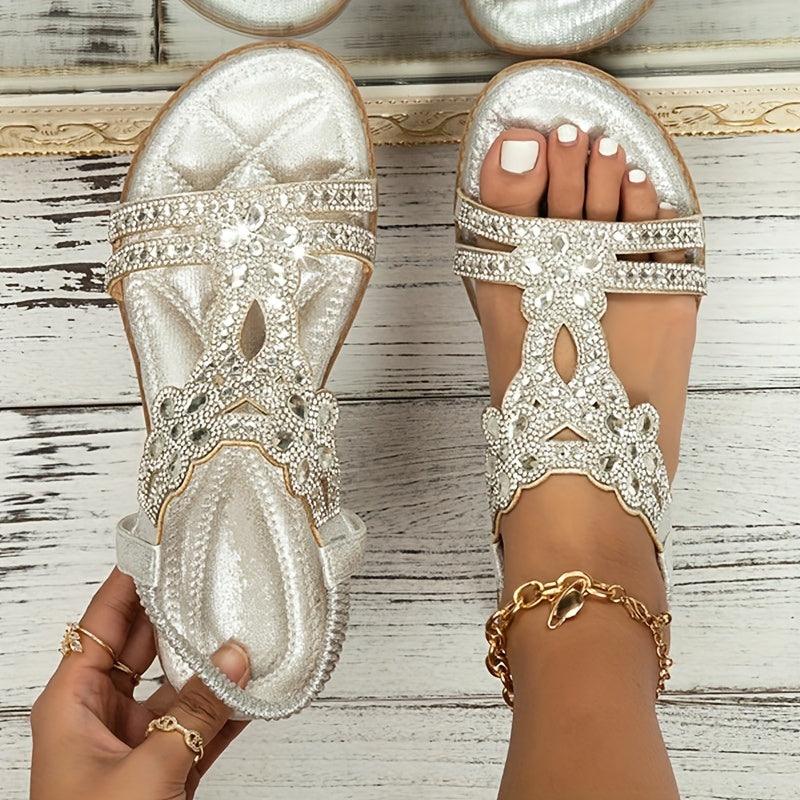 Silberne Sandalen mit Strassverzierung auf weißem Holzboden, Fuß mit goldener Fußkette und weiß lackierten Nägeln. Elegante Sommerschuhe Damen.