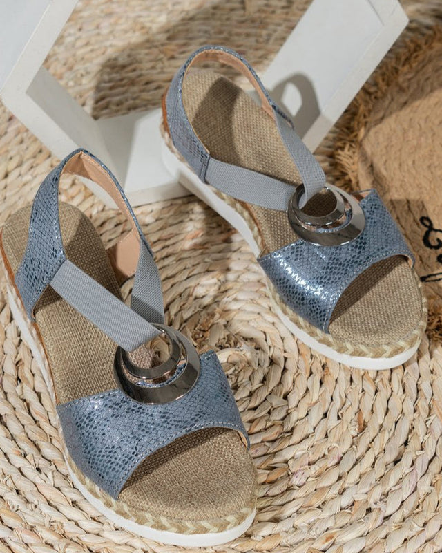 Silberne Sandalen mit elastischen Riemen und Metallringen auf geflochtener Unterlage, ideal für Sommermode und Strandoutfits.