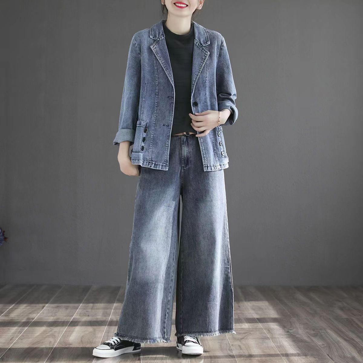 Frau in lässigem Denim-Outfit mit Jeansjacke und weiten Jeans, kombiniert mit schwarzen Sneakers, steht auf Holzboden vor grauem Hintergrund.