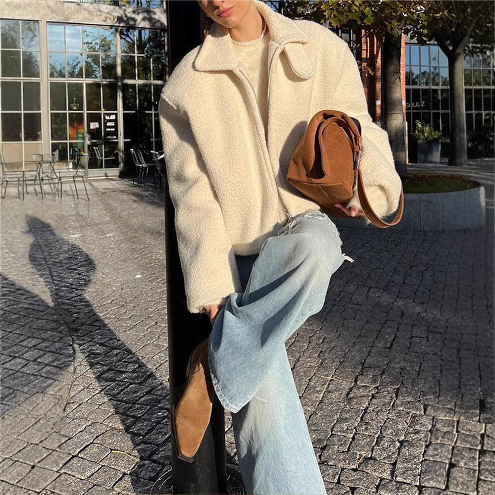 Person in weißer Teddyjacke und blauer Schlaghose hält braune Tasche, sitzt auf gepflastertem Platz vor modernen Glasgebäuden.