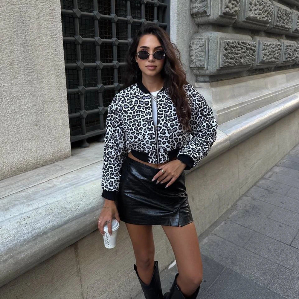 Frau in Leopardenjacke und schwarzem Lederrock, hält Kaffeebecher, steht vor historischer Steinwand, trägt Sonnenbrille und Stiefel.