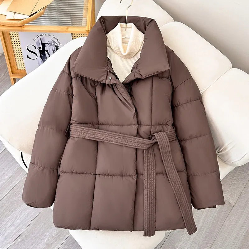 Braune Daunenjacke mit Gürtel auf weißem Sofa, modisch und warm, ideal für Wintermode. Perfekt für Damenbekleidung und Outdoor-Aktivitäten.