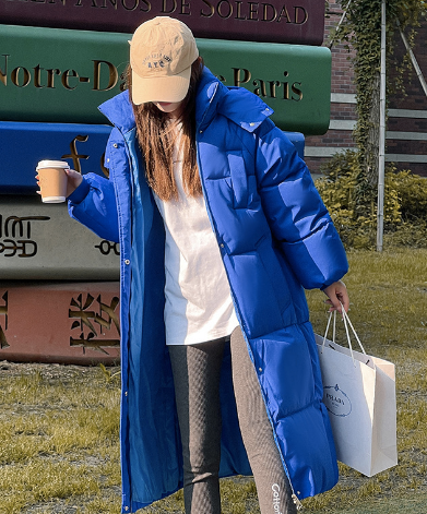 Frau in blauem Daunenmantel, hält Kaffeebecher und Einkaufstasche, steht vor bunten Schildern im Freien. Wintermode, Outdoor, Freizeitkleidung.