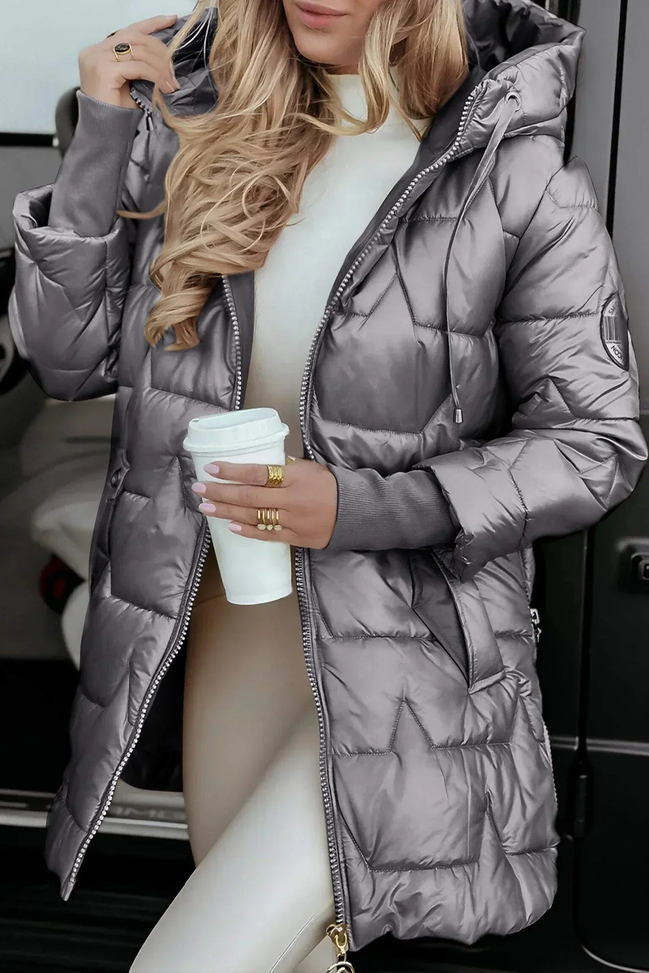 Frau in silberner Daunenjacke mit Kapuze hält Kaffeebecher, trägt beige Leggings. Wintermode, Outdoor-Bekleidung, stilvoller Look.