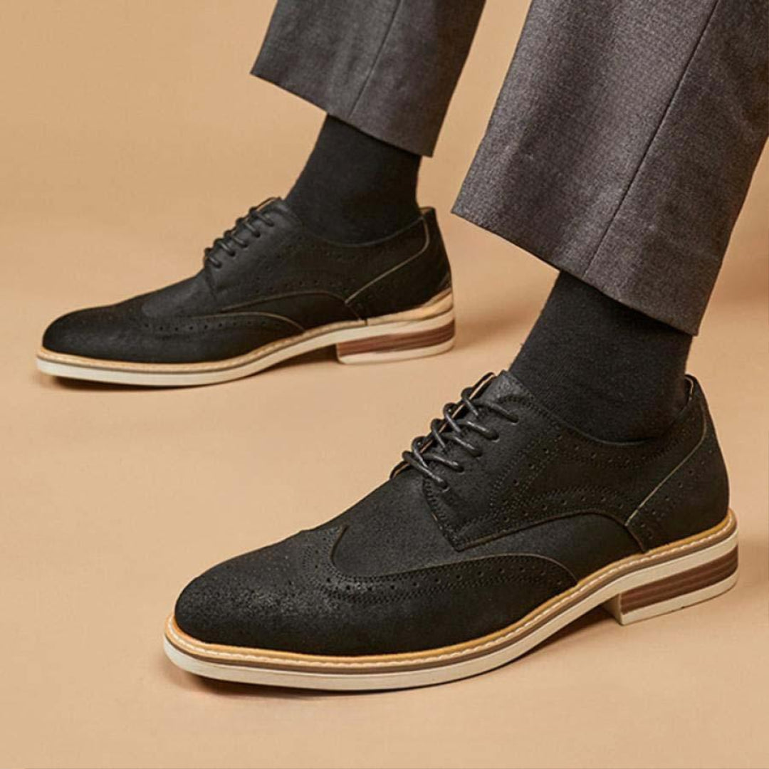 Schwarze Herren-Brogues aus Leder, getragen mit grauen Anzughosen und schwarzen Socken, auf beigem Hintergrund. Elegante Business-Schuhe.