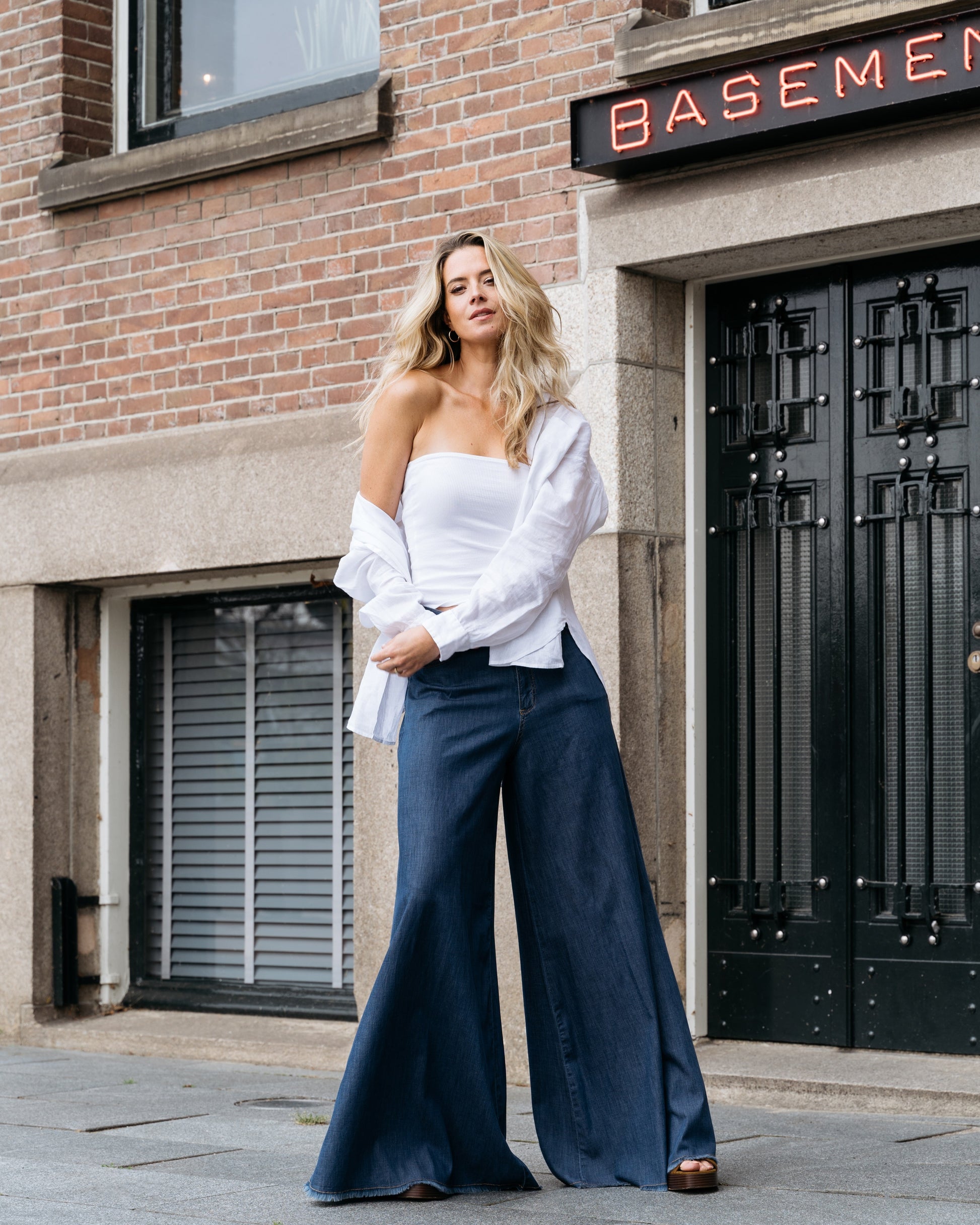 Frau in weißem Top und blauer Schlaghose vor Backsteingebäude mit "Basement"-Schild. Modetrend, Streetstyle, lässiger Look.