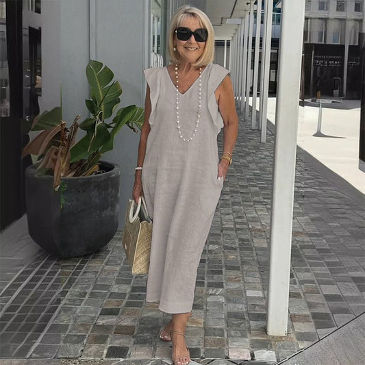 Ältere Frau in grauem Leinenkleid mit Perlenkette, Sonnenbrille und Strohtasche, steht lächelnd auf gepflastertem Gehweg neben Pflanze.