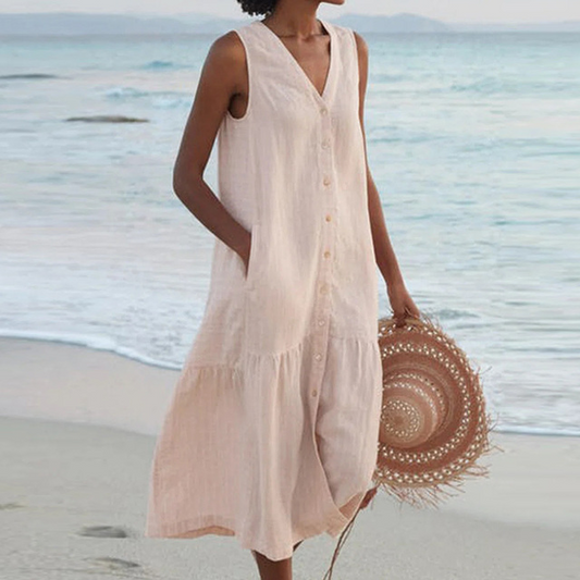 Frau in ärmellosem, beigem Sommerkleid am Strand, hält runden Strohhut. Meer im Hintergrund. Sommermode, Strandkleid, Strohhut.