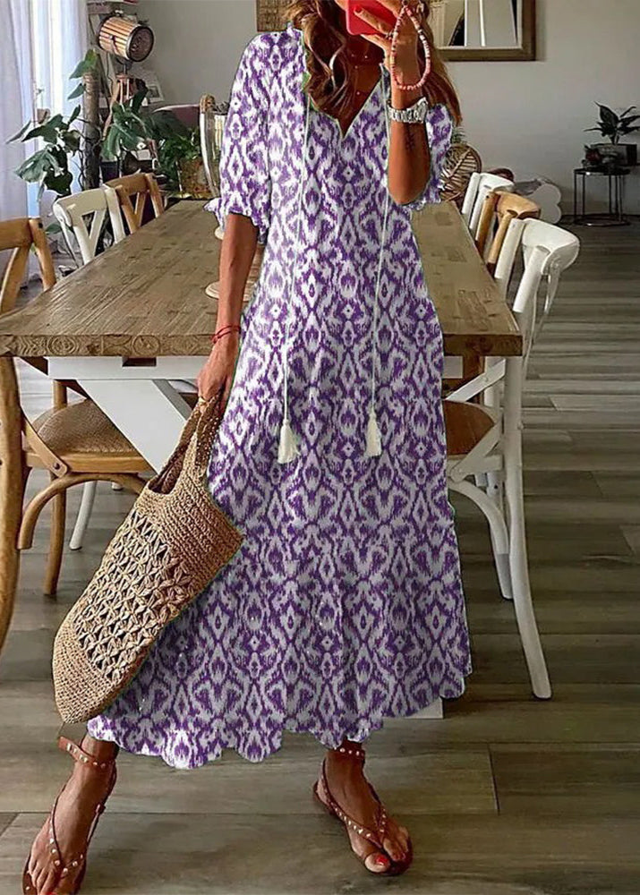 Frau in lila-weißem Boho-Kleid mit Ikat-Muster, hält eine geflochtene Tasche, steht in modernem Esszimmer mit Holztisch und weißen Stühlen.