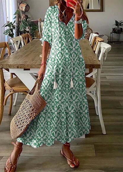 Frau in grün-weißem Ikat-Musterkleid, hält eine geflochtene Tasche, steht in modernem Esszimmer. Sommermode, Boho-Stil, Freizeitkleidung.