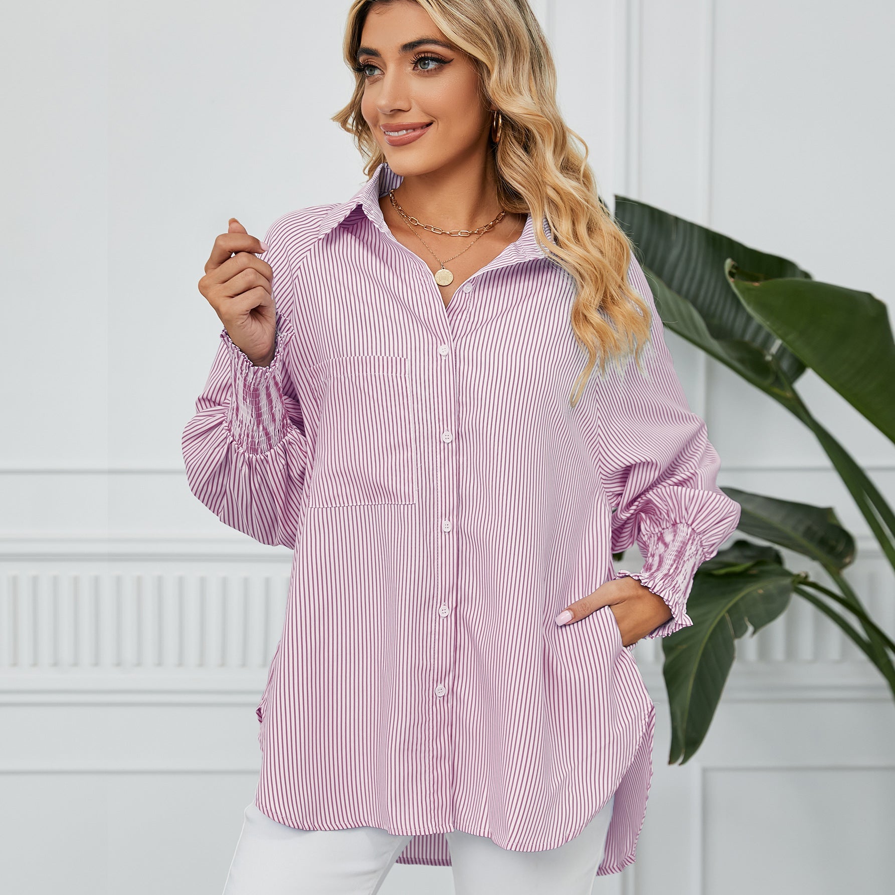 Frau in gestreifter Bluse, lächelnd, mit blonden Locken, steht vor weißer Wand mit Pflanze. Modetrend, Damenmode, lässiger Stil.