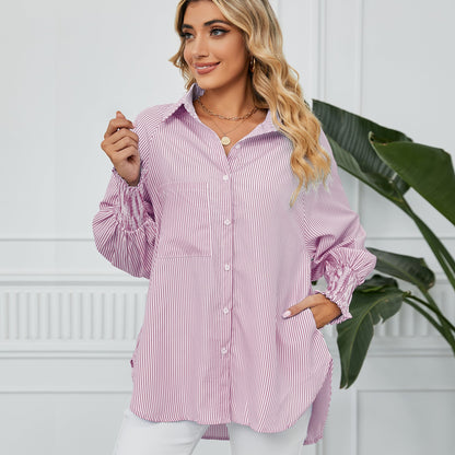 Frau in gestreifter Bluse, lächelnd, mit blonden Locken, steht vor weißer Wand mit Pflanze. Modetrend, Damenmode, lässiger Stil.