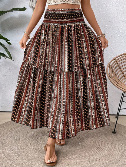 Frau trägt langen, gemusterten Boho-Rock in Rot- und Brauntönen, steht auf Teppich, neben Rattan-Stuhl und Pflanze. Boho-Mode, Sommerkleidung.