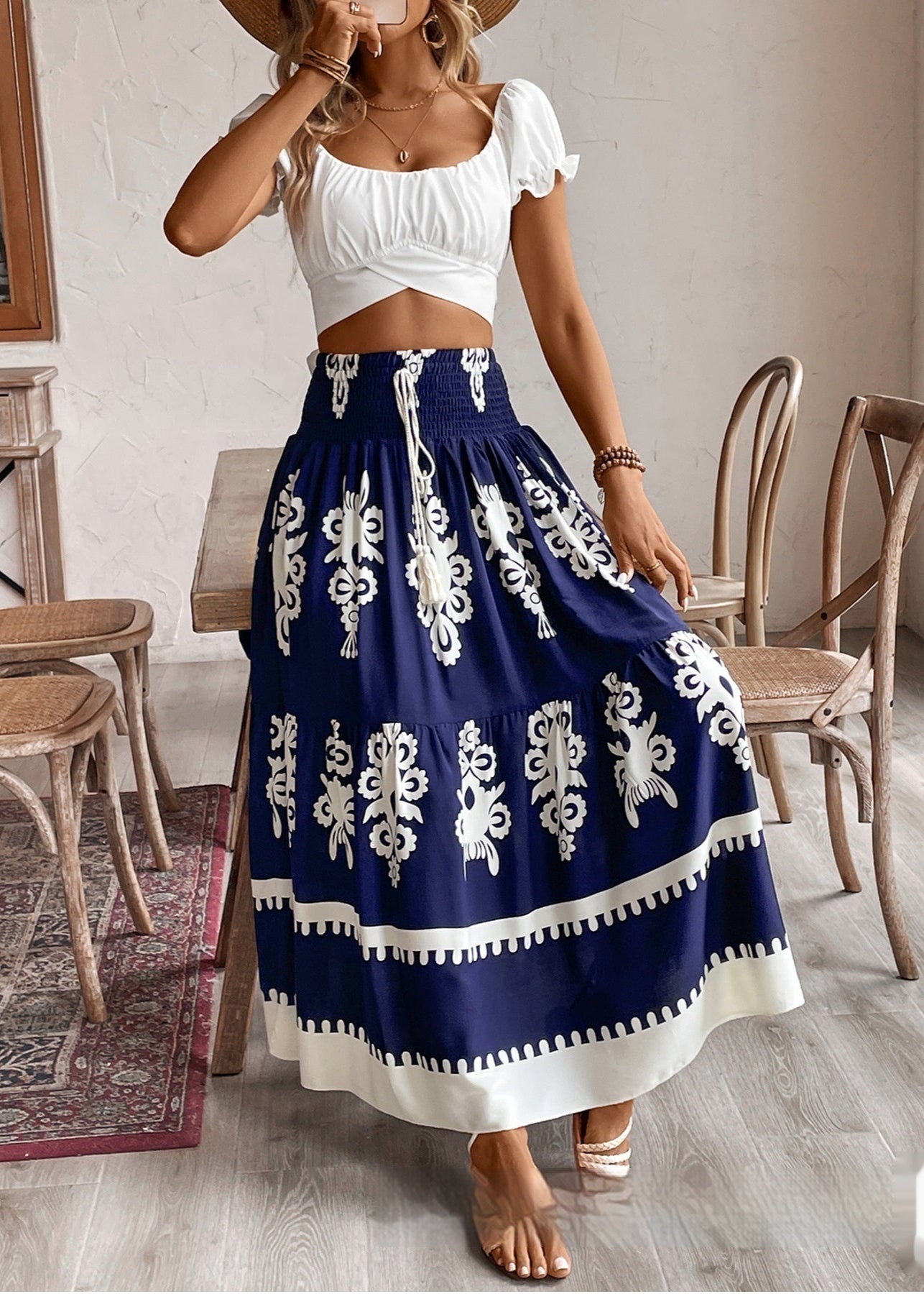Frau in weißem Crop-Top und blau-weißem Maxirock mit Blumenmuster, steht in modernem Café. Sommermode, Boho-Stil, trendige Damenbekleidung.